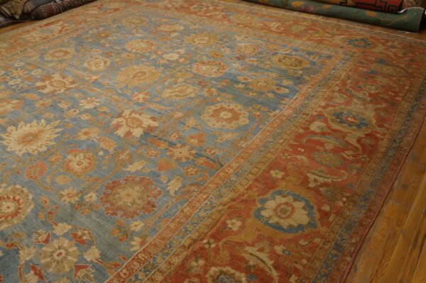 Muted blue floral rug with rust accents displayed on hardwood flooring next to rolled rugs.