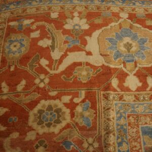 A traditional rug corner displays an elaborate floral pattern on a rust field, bordered by blue and beige bands.