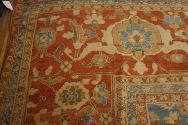 A traditional rug corner displays an elaborate floral pattern on a rust field, bordered by blue and beige bands.