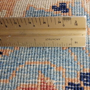 Detail view of rug texture and geometric pattern in blue, beige, and rust colors, measured by a ruler.
