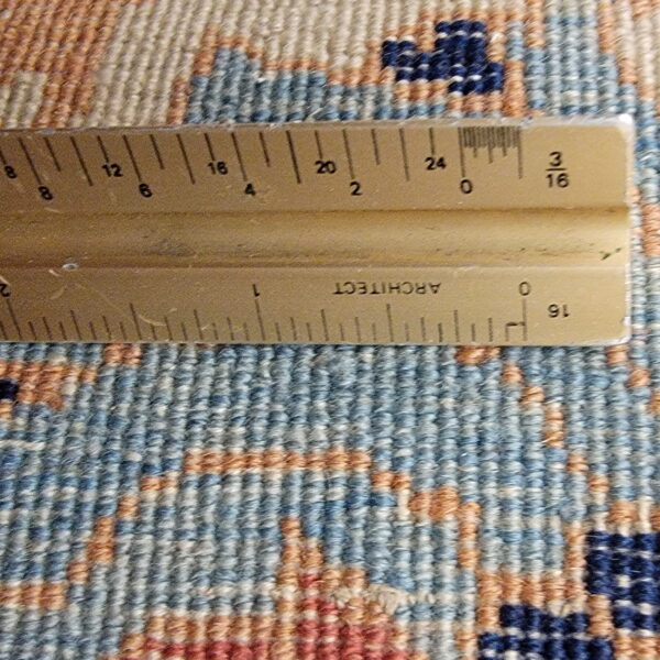 Detail view of rug texture and geometric pattern in blue, beige, and rust colors, measured by a ruler.