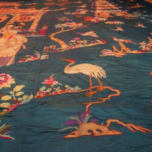 Detailed embroidery on a wrinkled teal textile featuring figures, pink flowers, and white cranes.