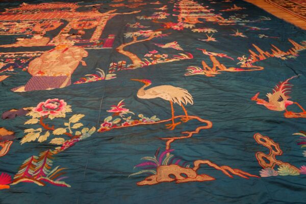 Detailed embroidery on a wrinkled teal textile featuring figures, pink flowers, and white cranes.