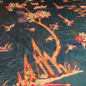 Close-up of a teal embroidered textile featuring bright pink flowers, tan trees, and flying birds.