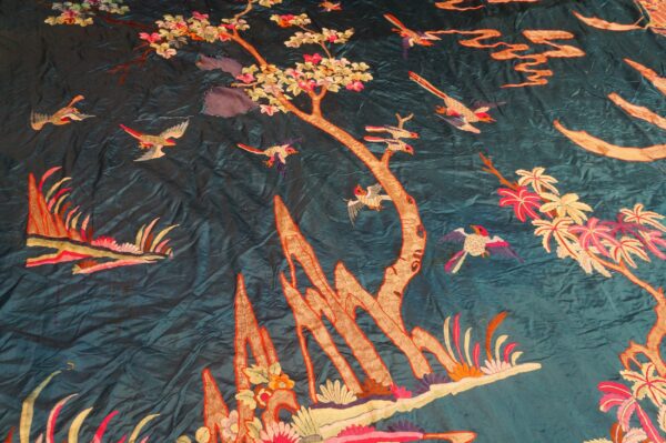 Close-up of a teal embroidered textile featuring bright pink flowers, tan trees, and flying birds.