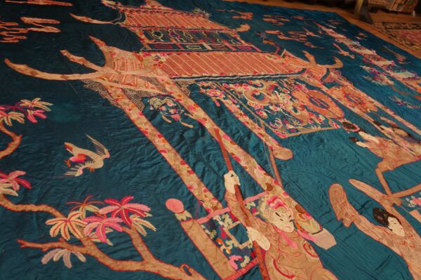 Teal textile with elaborate red, pink, and gold figures, a pagoda, and trees depicted across the surface.