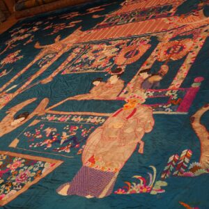 Teal embroidered flatweave rug with a detailed pictorial scene is spread out on a wooden floor.