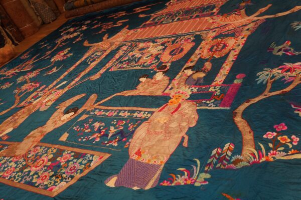 Teal embroidered flatweave rug with a detailed pictorial scene is spread out on a wooden floor.