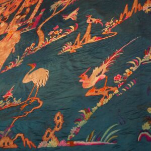 Wrinkled, glossy teal fabric embroidered with colorful birds, floral vines, and figures in a scenic pattern.