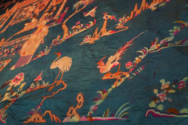 Wrinkled, glossy teal fabric embroidered with colorful birds, floral vines, and figures in a scenic pattern.