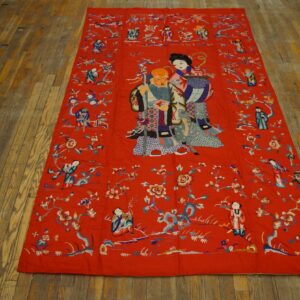 Red embroidered flatweave textile depicting figures and flowers rests on distressed wide plank flooring.