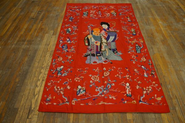 Red embroidered flatweave textile depicting figures and flowers rests on distressed wide plank flooring.