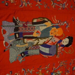 Detailed red textile rug featuring complex pictorial embroidery of figures and multicolored florals on wood planks.