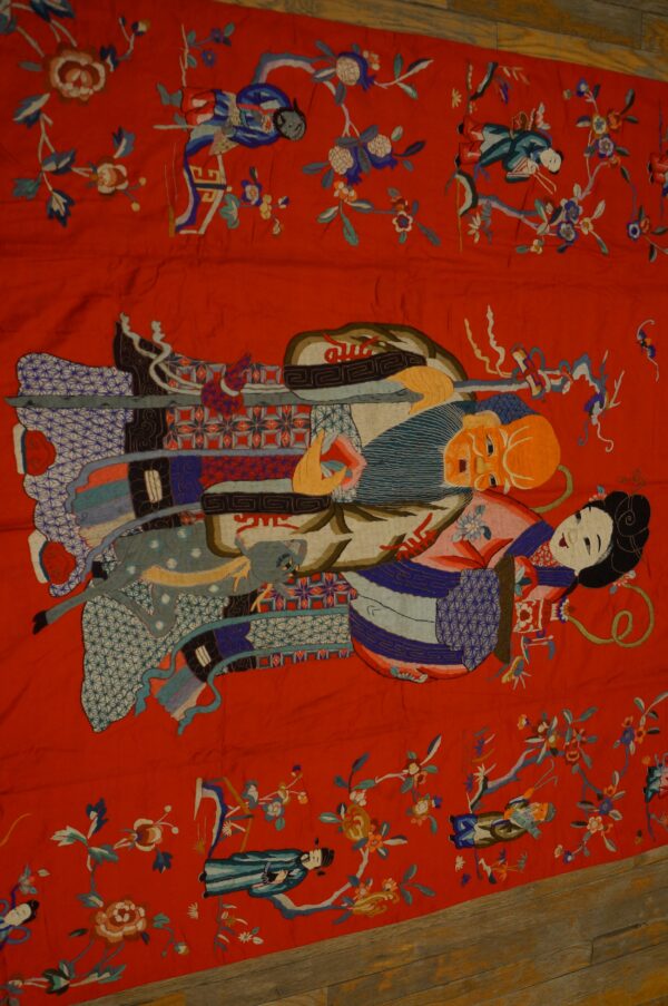 Detailed red textile rug featuring complex pictorial embroidery of figures and multicolored florals on wood planks.