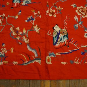 Low-pile red embroidered textile features blue, white, and brown floral and figural patterns over dark rustic wood flooring.