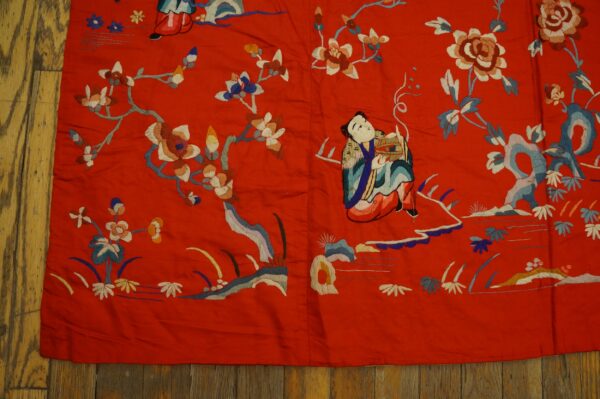 Low-pile red embroidered textile features blue, white, and brown floral and figural patterns over dark rustic wood flooring.