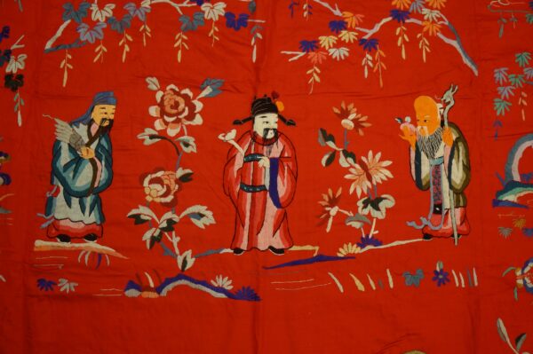 Detailed multicolored embroidery of three figures and flowers on a vibrant red, flat-pile surface.