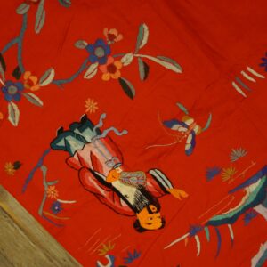 Vibrant red embroidered textile featuring floral and figurative motifs resting on rustic wood flooring.