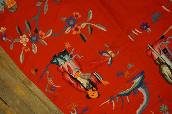 Vibrant red embroidered textile featuring floral and figurative motifs resting on rustic wood flooring.