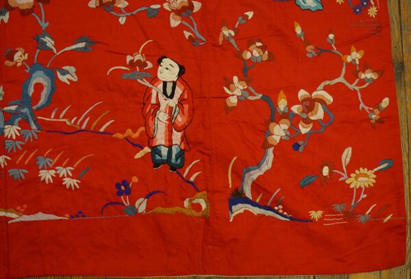 Scarlet flatweave textile embroidered with a central figure, multicolored flowers, and foliage on a wood floor.