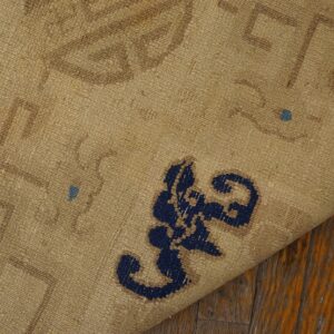 Tan rug backing with brown and blue patterns is folded over, exposing cream fringe on worn wood planks.
