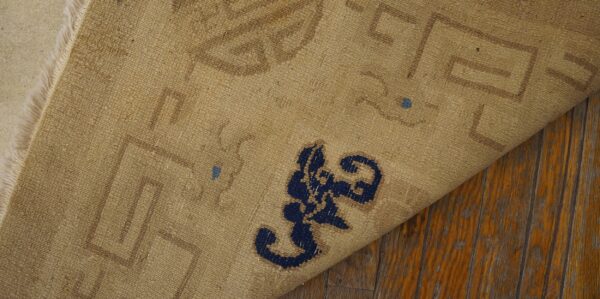 Tan rug backing with brown and blue patterns is folded over, exposing cream fringe on worn wood planks.