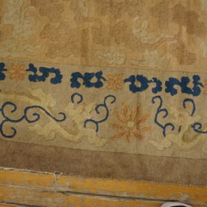 A detailed border of a gold and tan rug shows blue scrolls and orange flowers against distressed yellow wood flooring.