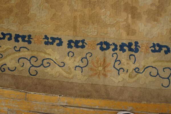 A detailed border of a gold and tan rug shows blue scrolls and orange flowers against distressed yellow wood flooring.