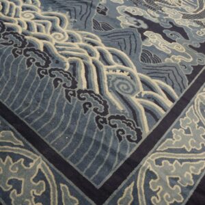 Blue low-pile rug with ornate waves, clouds, and border designs resting on wood flooring.