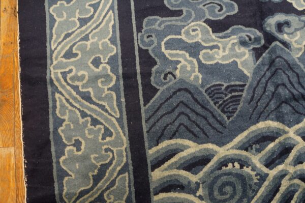 Dark blue rug with stylized light blue borders, mountains, and waves on a wooden floor.