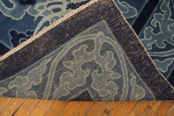 Dark blue ornate rug with scroll patterns is folded over, exposing its woven foundation on wood planks.