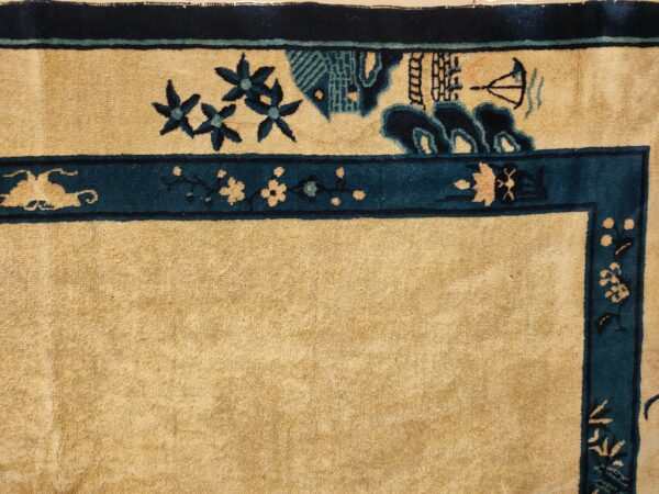 Tan rug detail with dark teal borders framing a field pattern of flowers, buildings, and a boat.