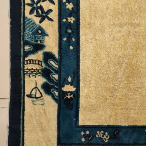 Gold rug field and thick pictorial teal border with house and boat designs on a light floor.