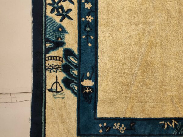 Gold rug field and thick pictorial teal border with house and boat designs on a light floor.