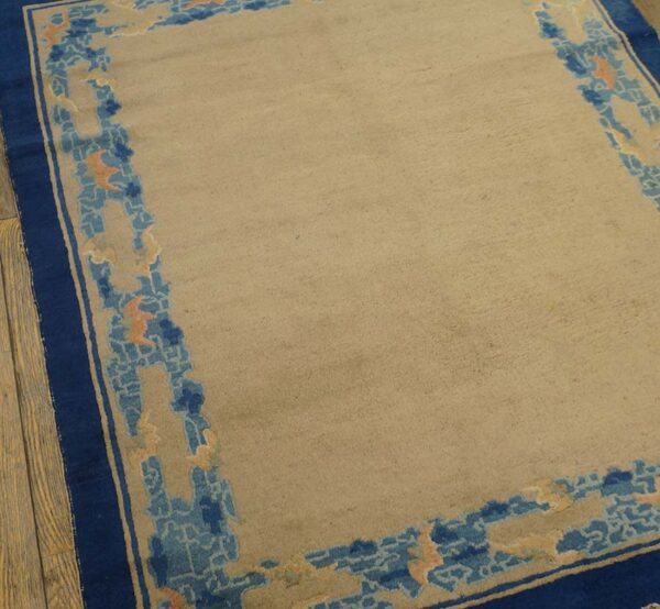 Tan rug with a wide navy blue border and abstract light blue and orange patterns, resting on wood planks.