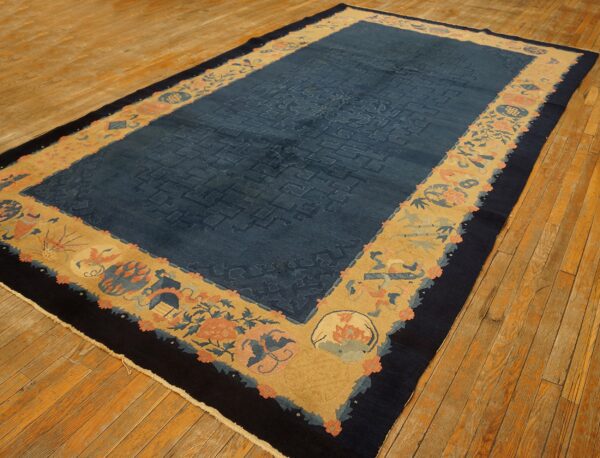 Indigo and gold rug featuring a subtle fretwork field and a wide floral-and-object border on wood flooring.