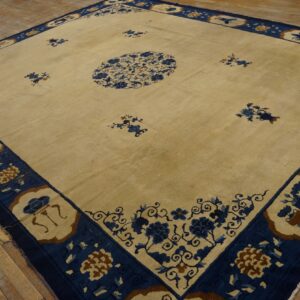 Beige square rug with an elaborate navy blue border and scattered designs on wood flooring.