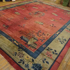 Red field rug with blue and gold floral motifs, wide olive border, displayed on distressed wood planks.