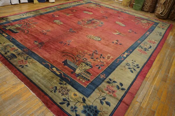 Red field rug with blue and gold floral motifs, wide olive border, displayed on distressed wood planks.