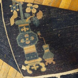 A rolled dark blue rug with tan and teal vase motifs rests on wide plank wood floors.