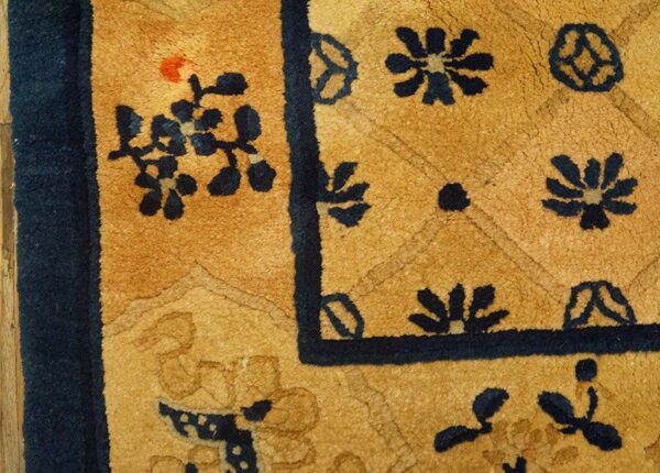Golden tan rug corner closeup with navy blue floral designs and a wide outer border near wood flooring.