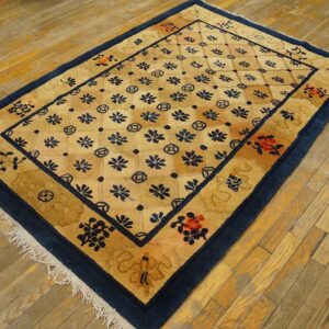 A rectangular golden tan rug with a navy blue floral lattice pattern and wide border sits on worn hardwood floors.