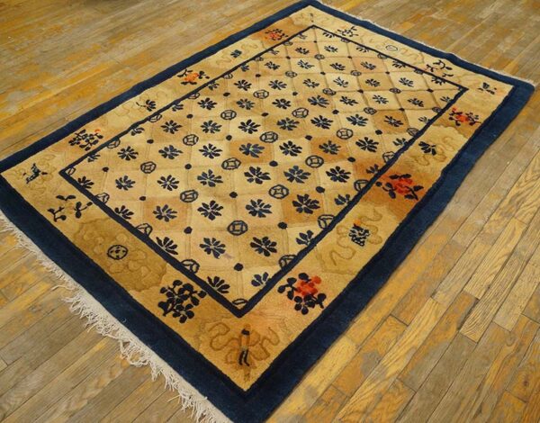A rectangular golden tan rug with a navy blue floral lattice pattern and wide border sits on worn hardwood floors.