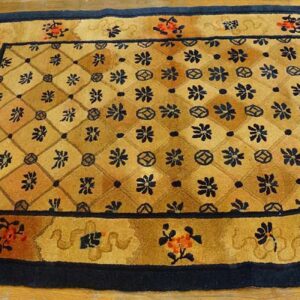 Tan and gold floral lattice rug with navy borders and fringe, placed on light wooden plank flooring.