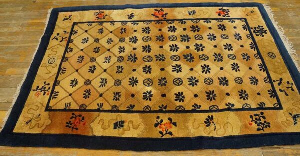 Tan and gold floral lattice rug with navy borders and fringe, placed on light wooden plank flooring.