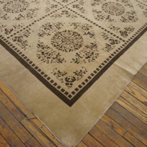 Corner view of a low-pile beige rug with dark brown diamond patterns on worn wooden plank flooring.