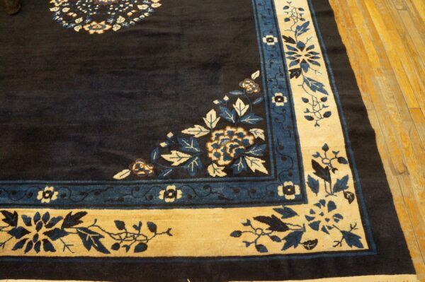 Close-up of a navy rug corner showing tan and blue floral borders on light wood flooring.