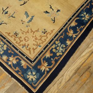 Close-up of a tan rug with blue floral patterns and a wide navy border on light wooden floorboards.