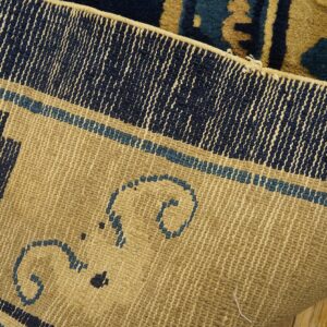 A rug's woven khaki backing, displaying navy and blue border details, rests on wooden planks.