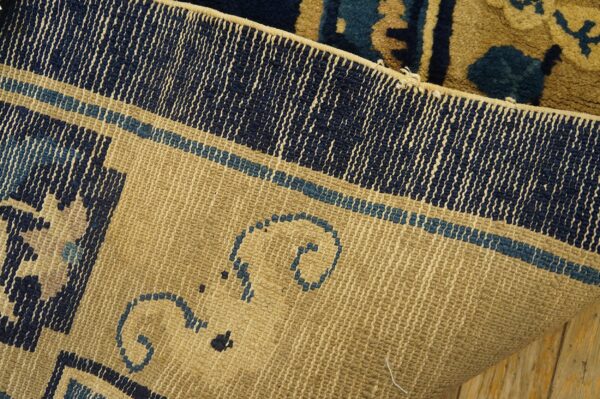 A rug's woven khaki backing, displaying navy and blue border details, rests on wooden planks.